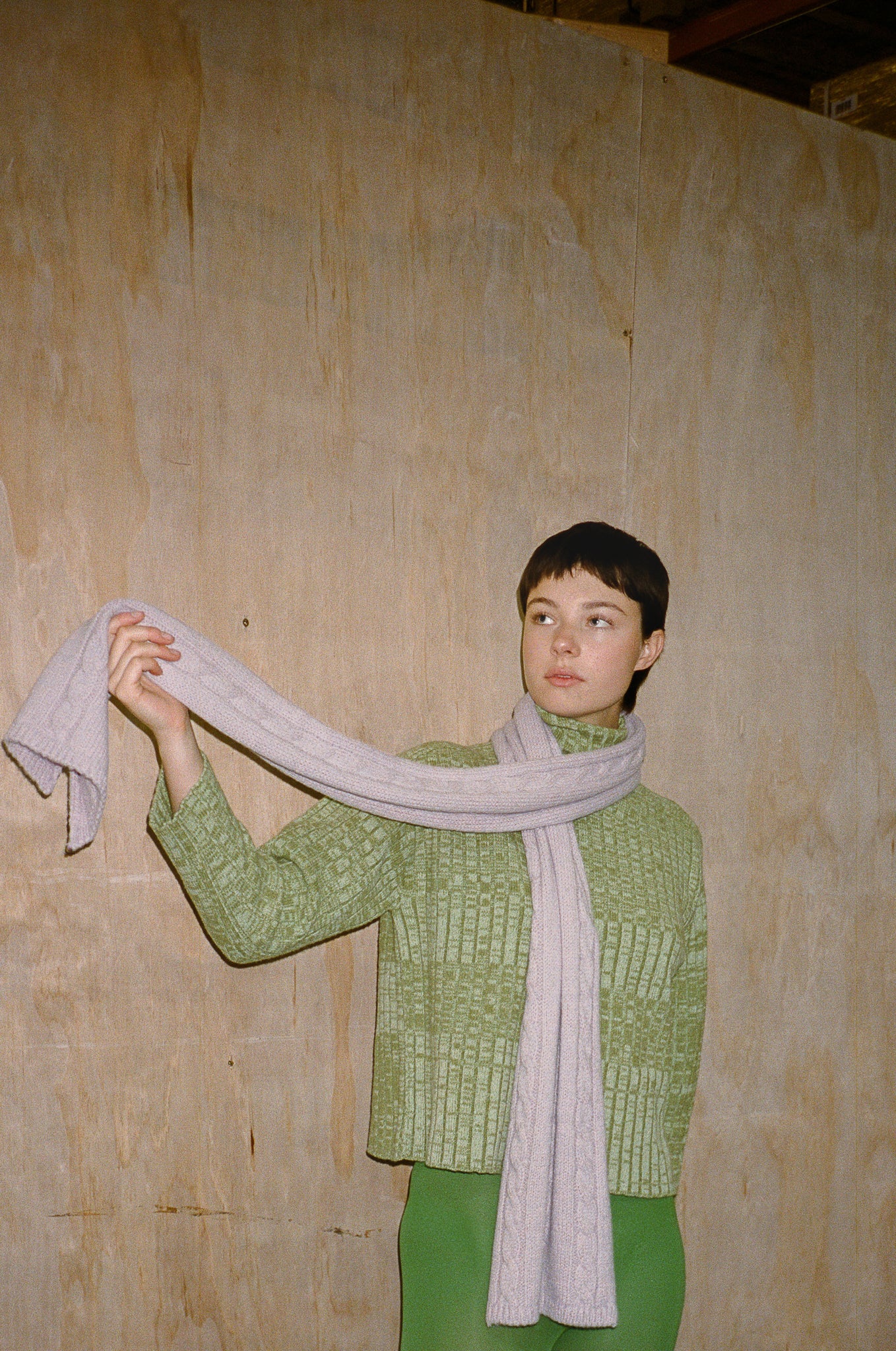 Person wearing a green knitted sweater and holding a light gray scarf against a wooden wall.