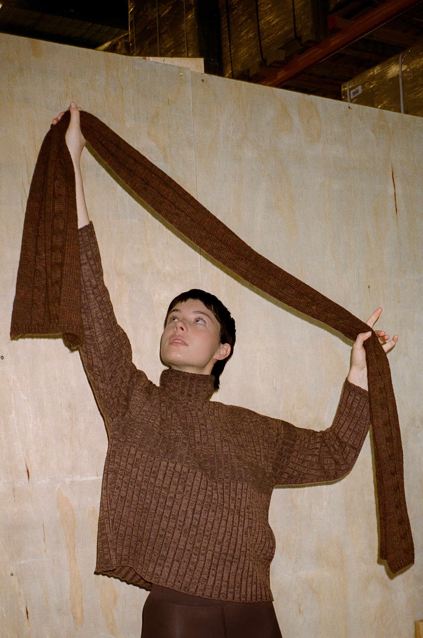 Person wearing a brown sweater holding a long brown fabric against a plain wall.