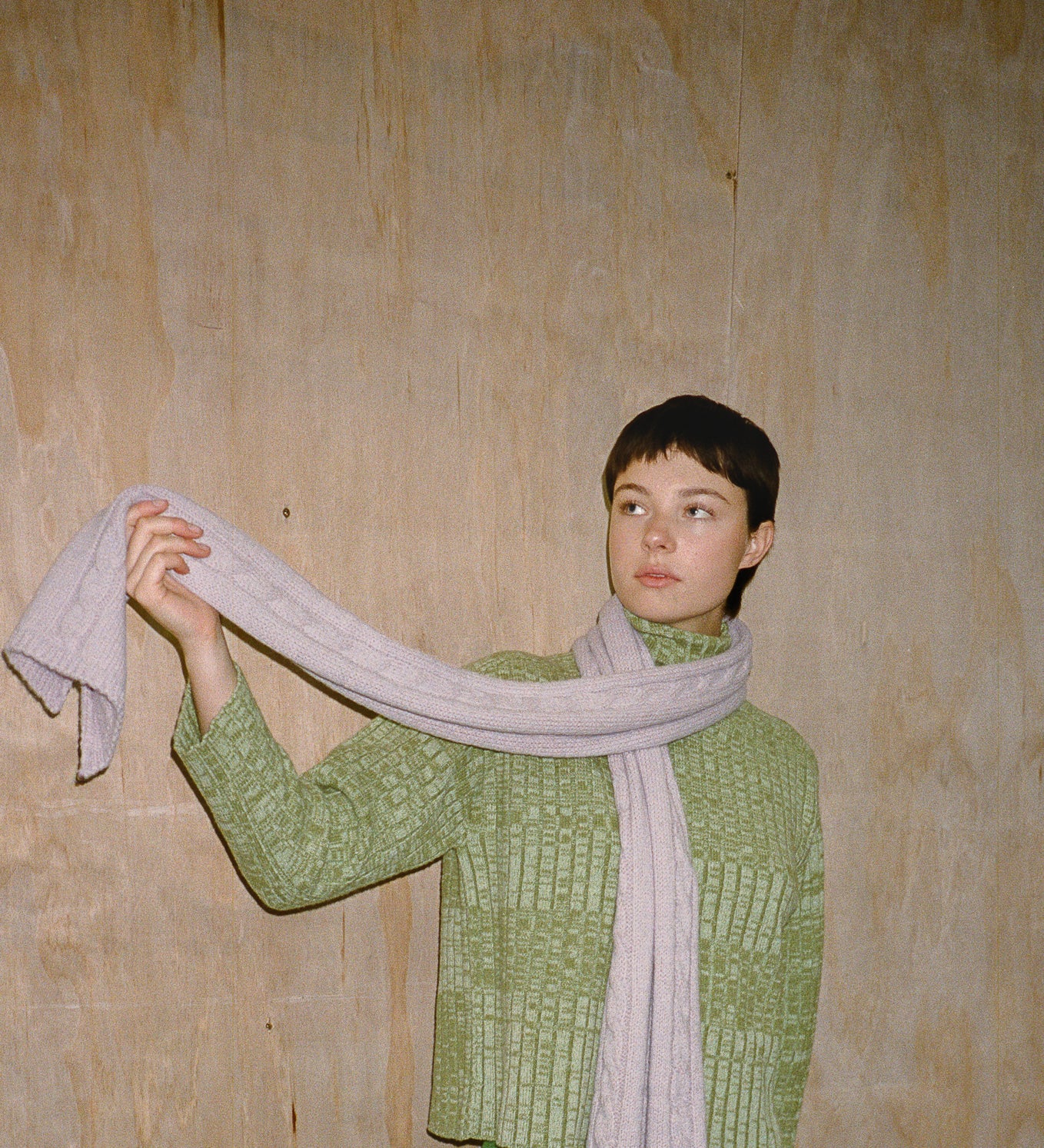 Person wearing a green knitted sweater and holding a light gray scarf against a wooden wall.