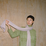 Person wearing a green knitted sweater and holding a light gray scarf against a wooden wall.