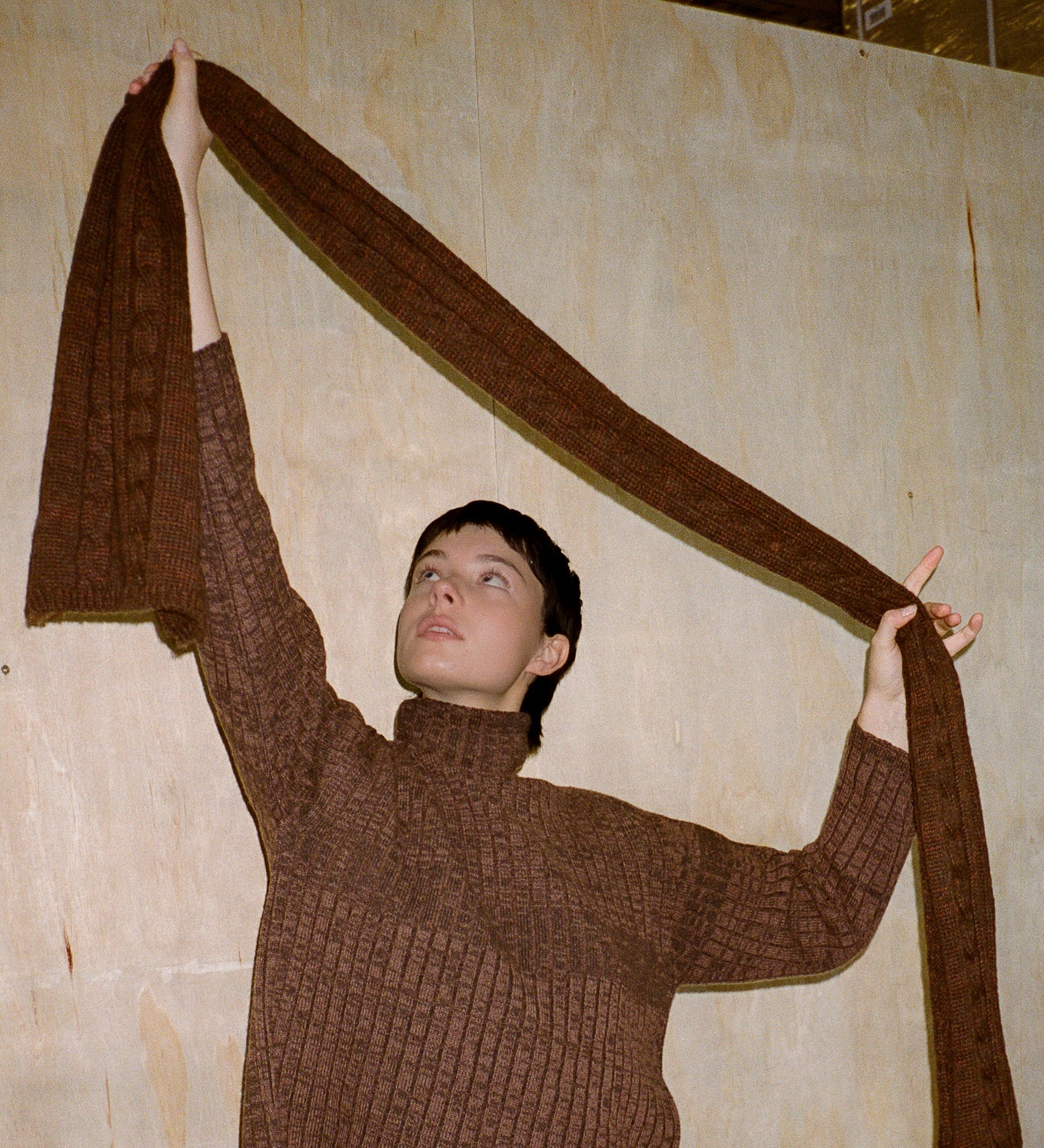 Person wearing a brown sweater holding a long brown fabric against a plain wall.