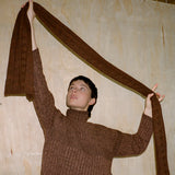 Person wearing a brown sweater holding a long brown fabric against a plain wall.