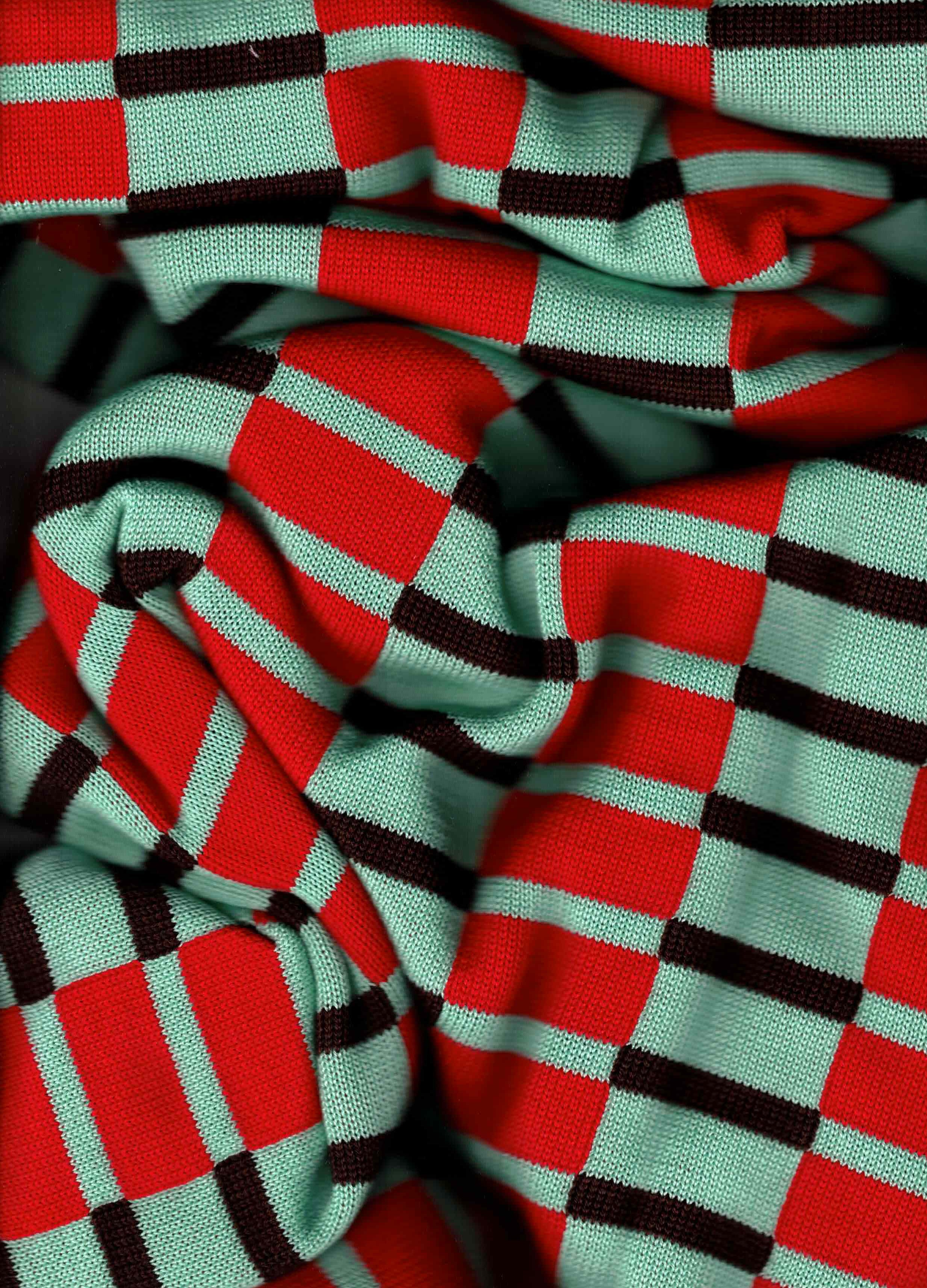 A close-up of a knitted fabric with a red, mint green, and black checkered pattern, shown in soft folds. Australian made 